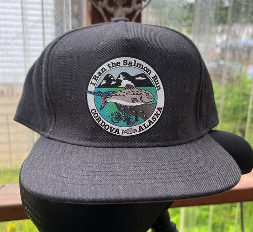 A dark gray heathered flat-brim cap displayed on a black mannequin head, featuring a circular embroidered patch on the front with a salmon, mountains, and the text 'I Ran the Salmon Run Cordova Alaska'.